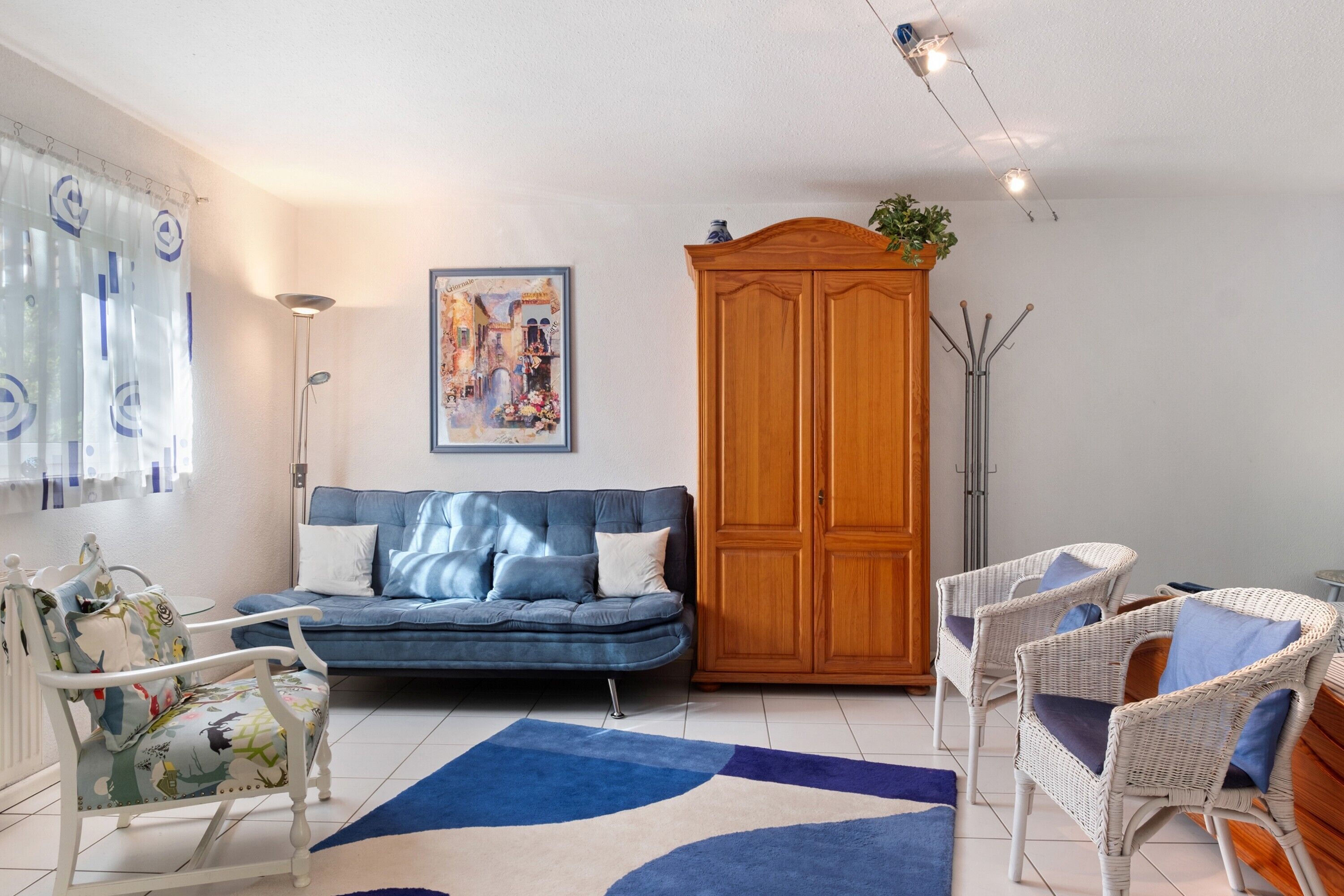 Ein Wohnzimmer mit einem blauen Sofa, weißen Korbstühlen, einer Holzkommode und einem Gemälde an der Wand.