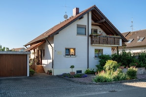 A two-story building with a sloped roof, a balcony with wooden railings, and a small garden with potted plants and flowers.