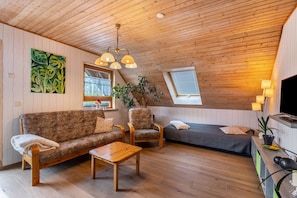 A cozy room with a wooden ceiling, a sofa, a chair, a small table, a bed, and a TV.