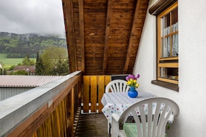A balcony with a wooden ceiling, a white chair, a table with a polka dot tablecloth, and a vase with flowers.