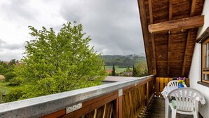 Una terrassa de fusta amb una cadira i taula blanca, amb vista a un paisatge verd amb arbres i cases a la distància.