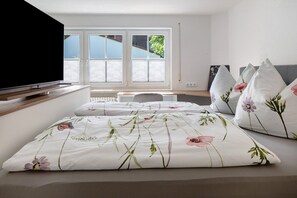 Een slaapkamer met een bed, kussens, een televisie, een bureau en een raam met jaloezieën.