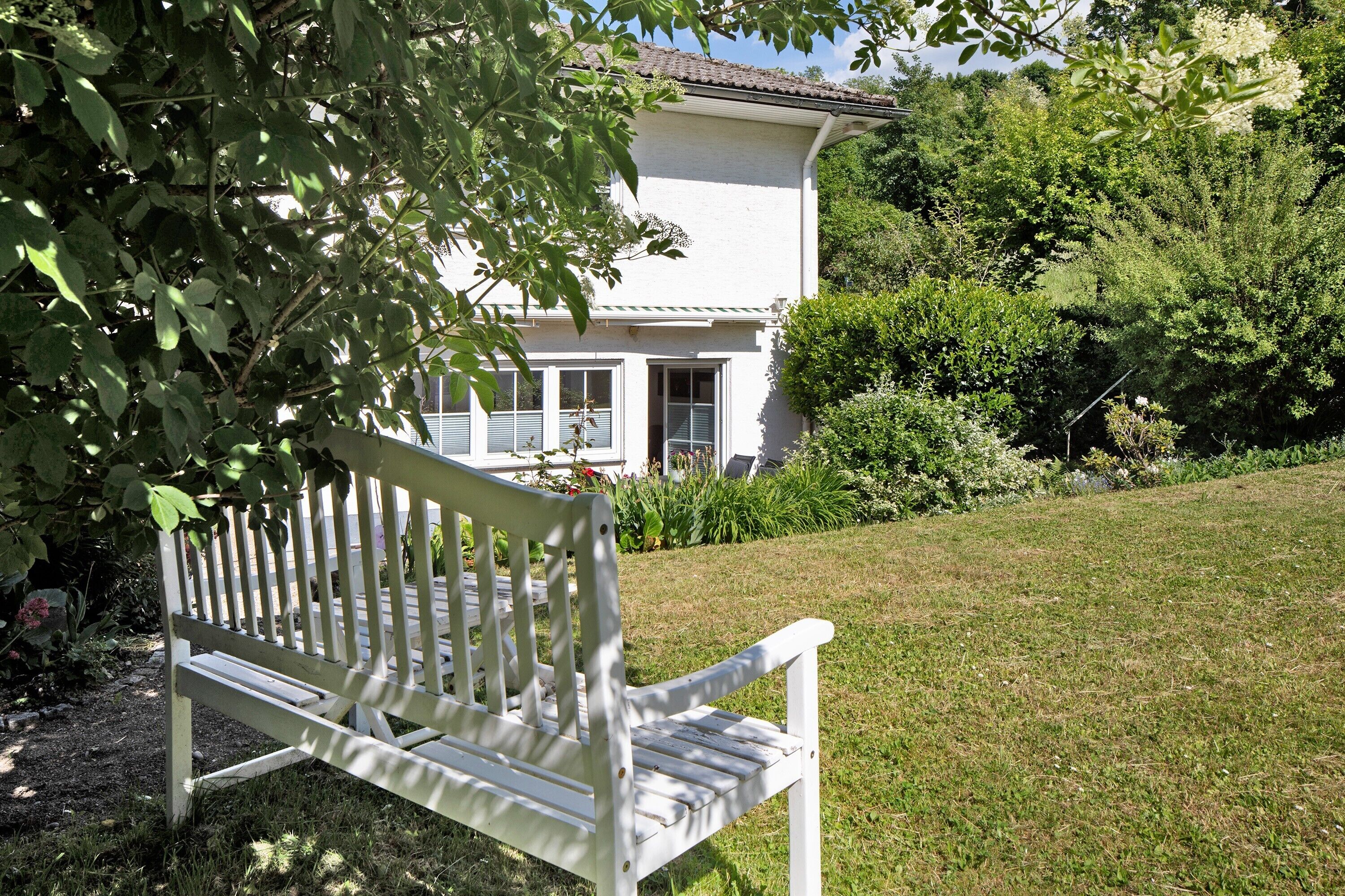 Eine weiße Bank in einem Garten mit einem weißen Gebäude im Hintergrund.