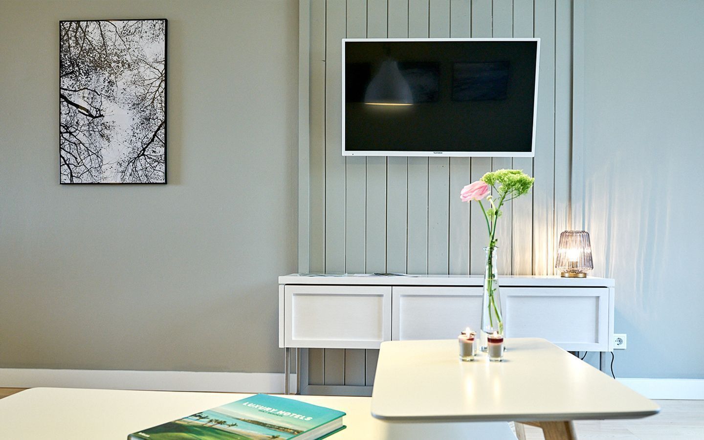 Moderni hotellihuone, jossa on seinälle asennettu taulutelevisio, valkoinen konsolipöytä, vaasi, jossa on vaaleanpunainen kukka, ja pöydällä kirja.