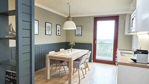 A modern kitchen with a wooden dining table, a hanging light fixture, and a view of the outdoors through a large window.