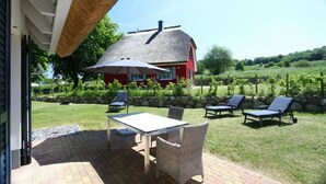Een terras met een tafel en stoelen, een rieten dak en een rode woning op de achtergrond.