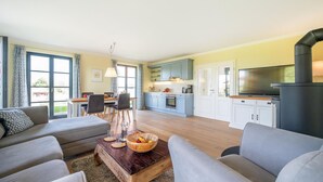 Een moderne woonkamer met een bank, salontafel en een eethoek met stoelen en een tafel. Achtergrond toont een keuken met kasten en apparatuur.