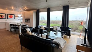 A spacious living area with a large black sofa, a glass coffee table, and a dining area with a white table and chairs. - Holiday Home 'Großzügiges Modernes' with Shared Terrace, Private Garden and Wi-Fi (Neuharlingersiel)