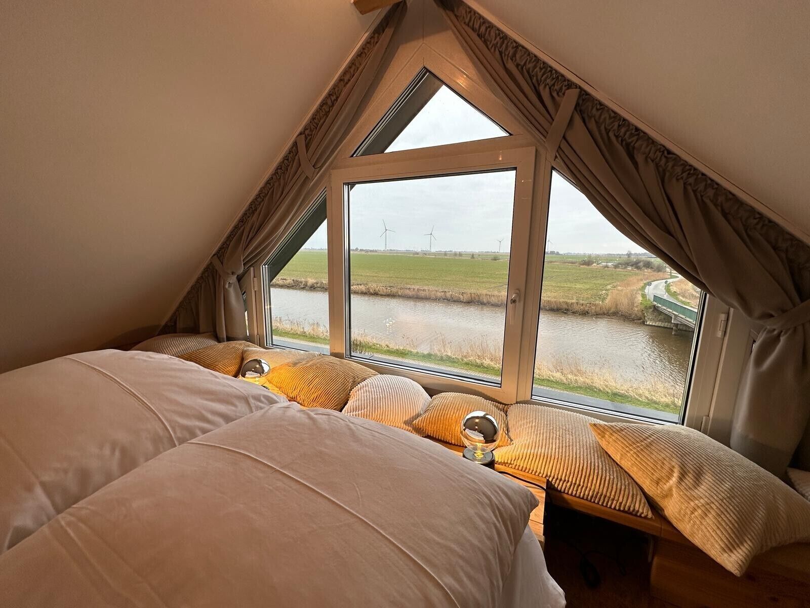 Un dormitorio con un ventanal grande, una cama y vistas a un canal y aerogeneradores.