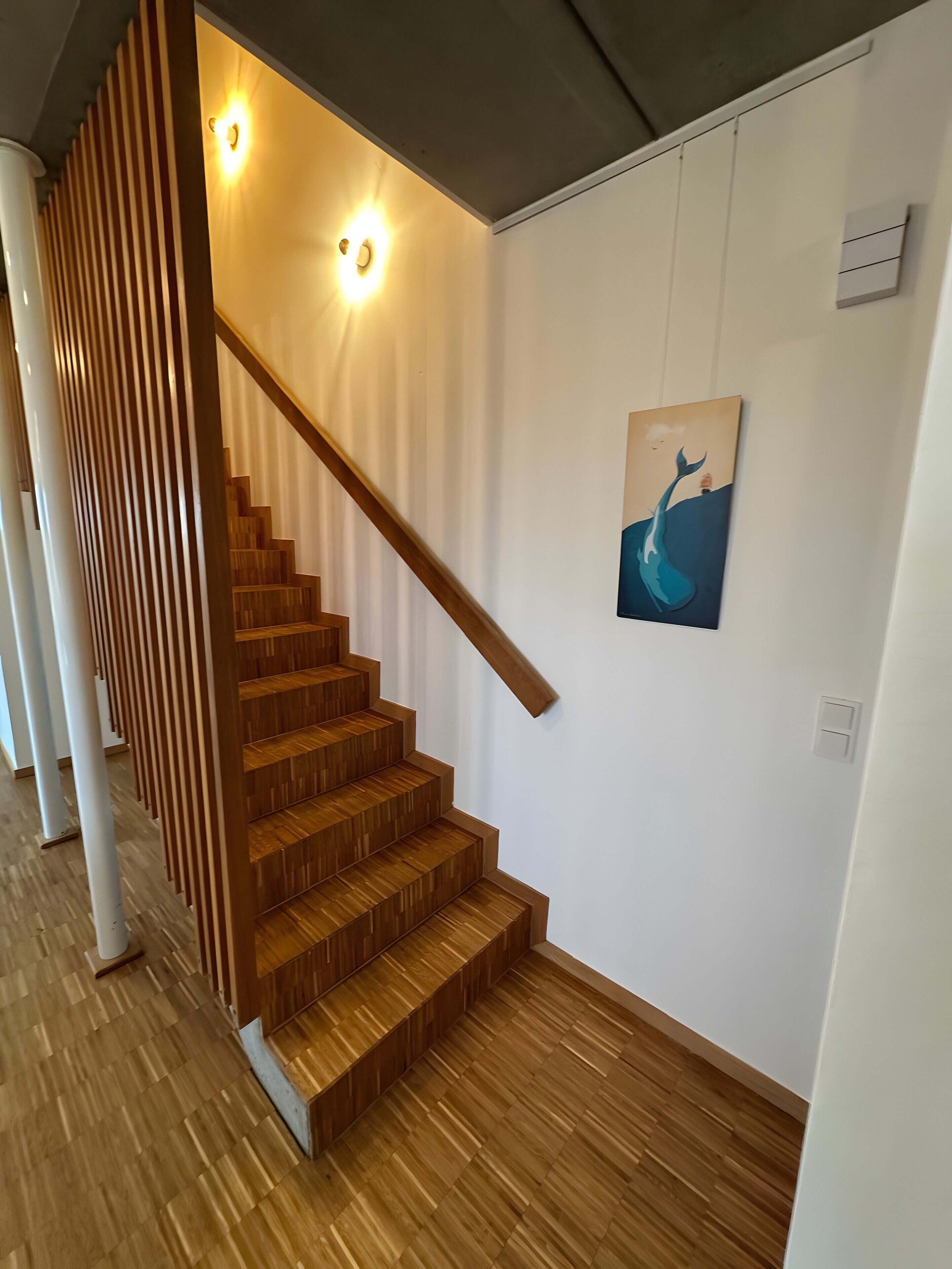 Un escalier avec des marches en bois et une rampe en bois, menant à un palier où se trouve un luminaire fixé au mur et un tableau représentant une baleine.