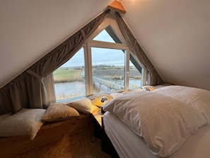 A bedroom with a sloped ceiling, large windows, a wooden desk, and a bed with white bedding.