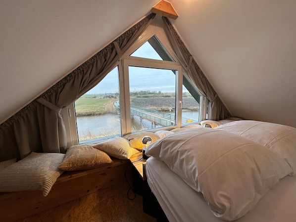 A bedroom with a sloped ceiling, large windows, a wooden desk, and a bed with white bedding.