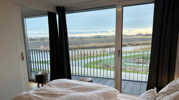 A bedroom with a large bed, a view of a river, and a balcony with a railing. - Holiday Home 'Großzügiges Modernes' with Shared Terrace, Private Garden and Wi-Fi (Neuharlingersiel)