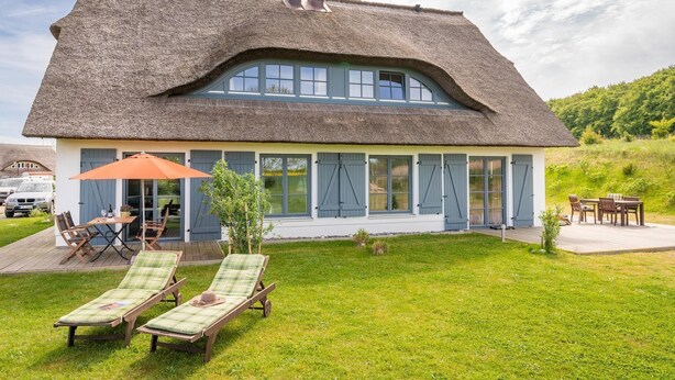 Ein Haus mit einem Reetdach, einer großen Terrasse, zwei Liegestühlen und einem Tisch mit Sonnenschirm.