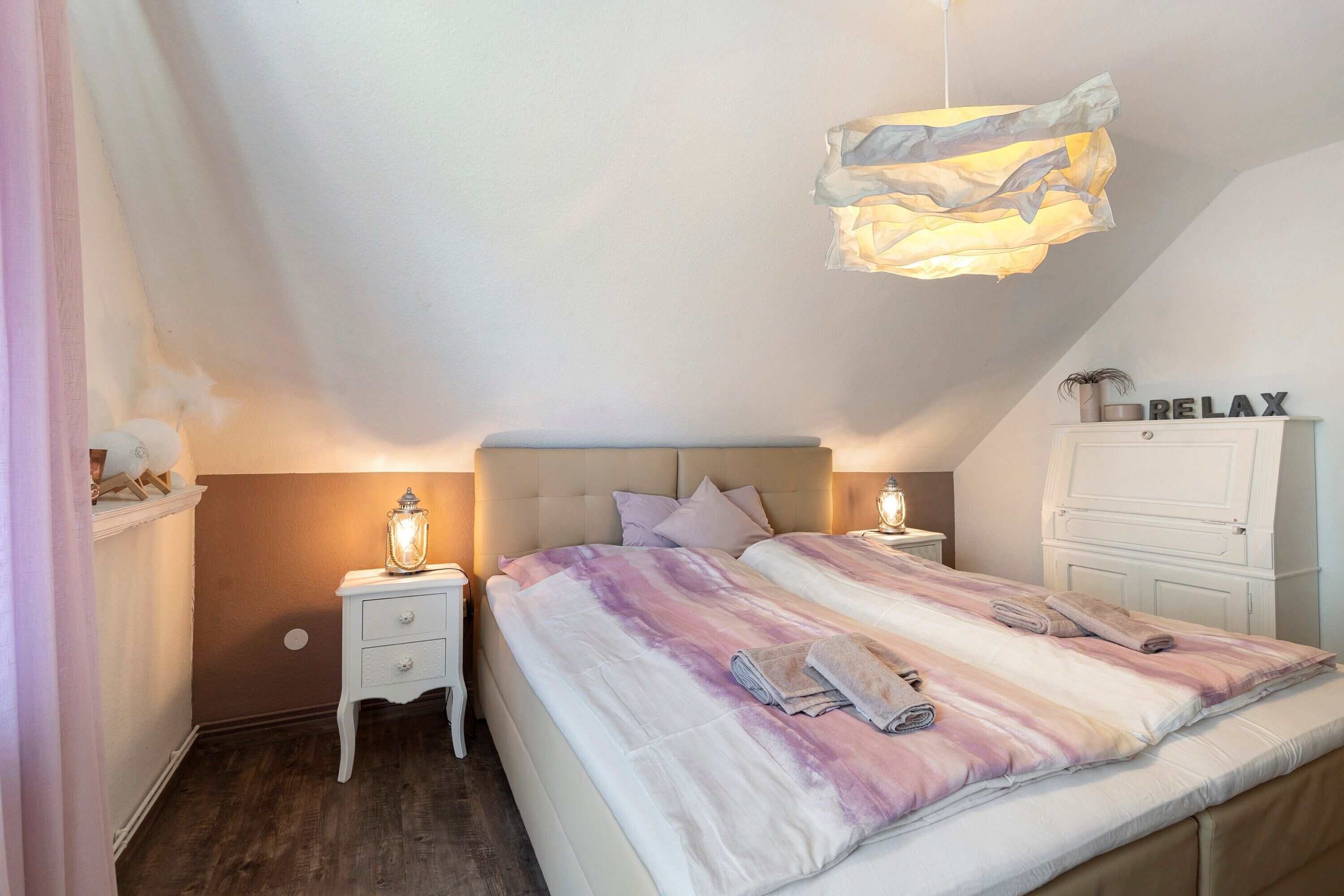 Een slaapkamer met een bed, nachtkastjes, een hanglamp en een dressoir met een bordje.