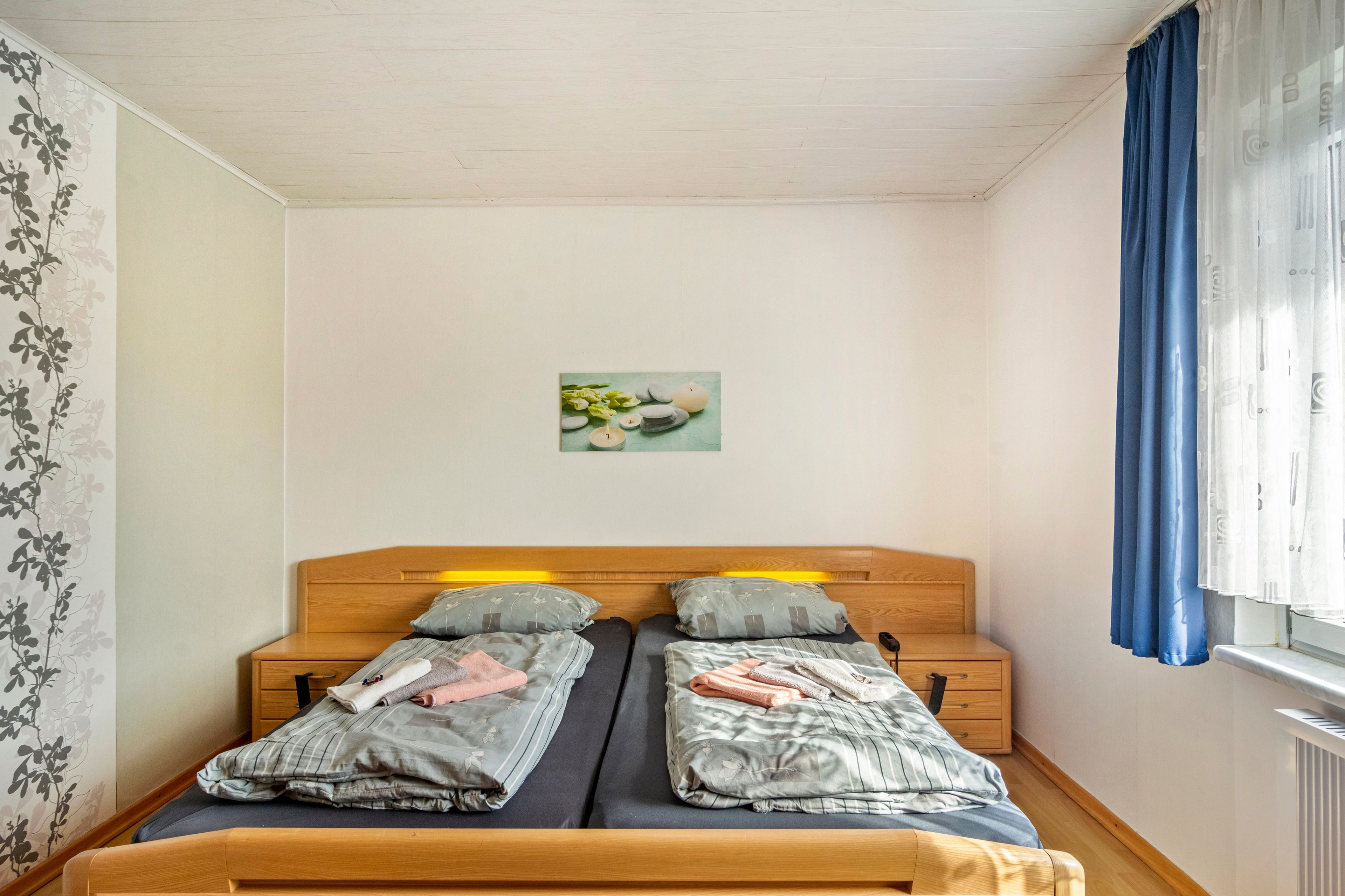 A double bed with two pillows, a bedside table, a small drawer, and a wall-mounted picture.