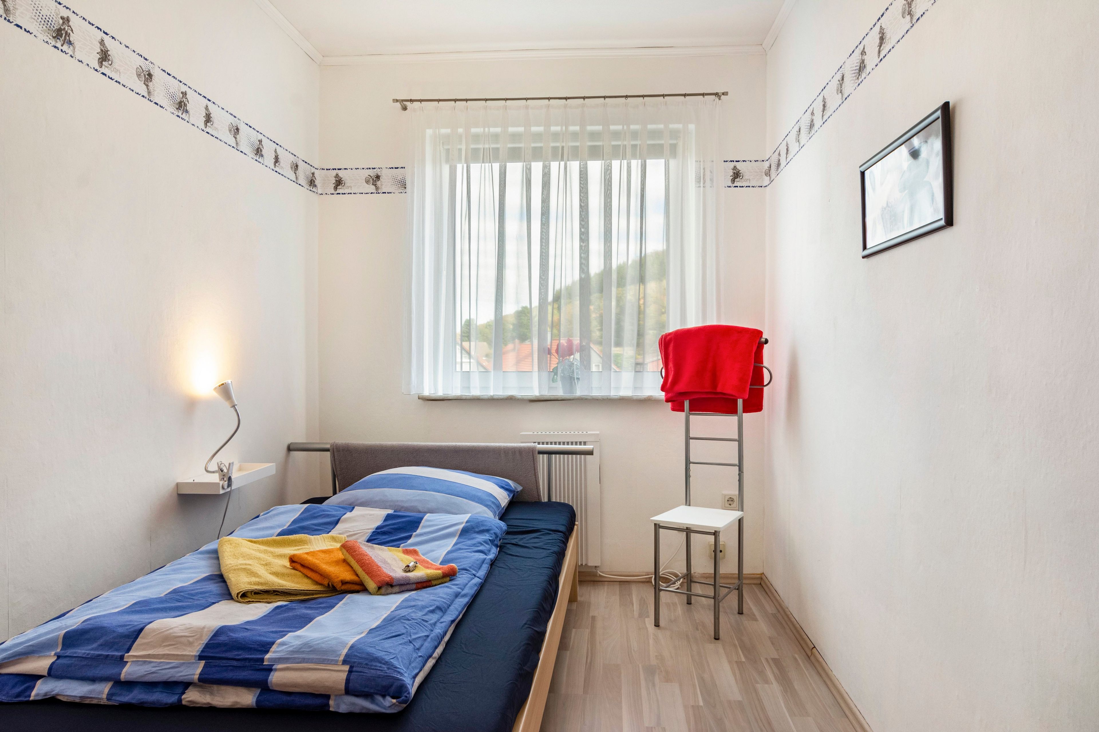 Ein kleines Zimmer mit einem Einzelbett, einem Fenster, einer Heizung, einem kleinen Tisch und einem roten Stuhl.
