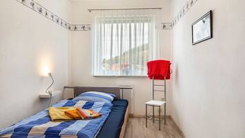 Ein kleines Zimmer mit einem Einzelbett, einem Fenster, einer Heizung, einem kleinen Tisch und einem roten Stuhl.