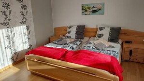 A double bed with a red blanket, two bedside tables, a nightstand, a wall-mounted painting, and a window with floral curtains. - Apartment 'Wolfswind' with Mountain View, Balcony and Wi-Fi (Langelsheim)