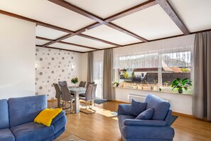 A modern living room with a dining area, featuring a large window with a view of the neighborhood, a blue sofa with a yellow pillow, and a dining table with chairs. - Apartment 'Wolfswind' with Mountain View, Balcony and Wi-Fi (Langelsheim)