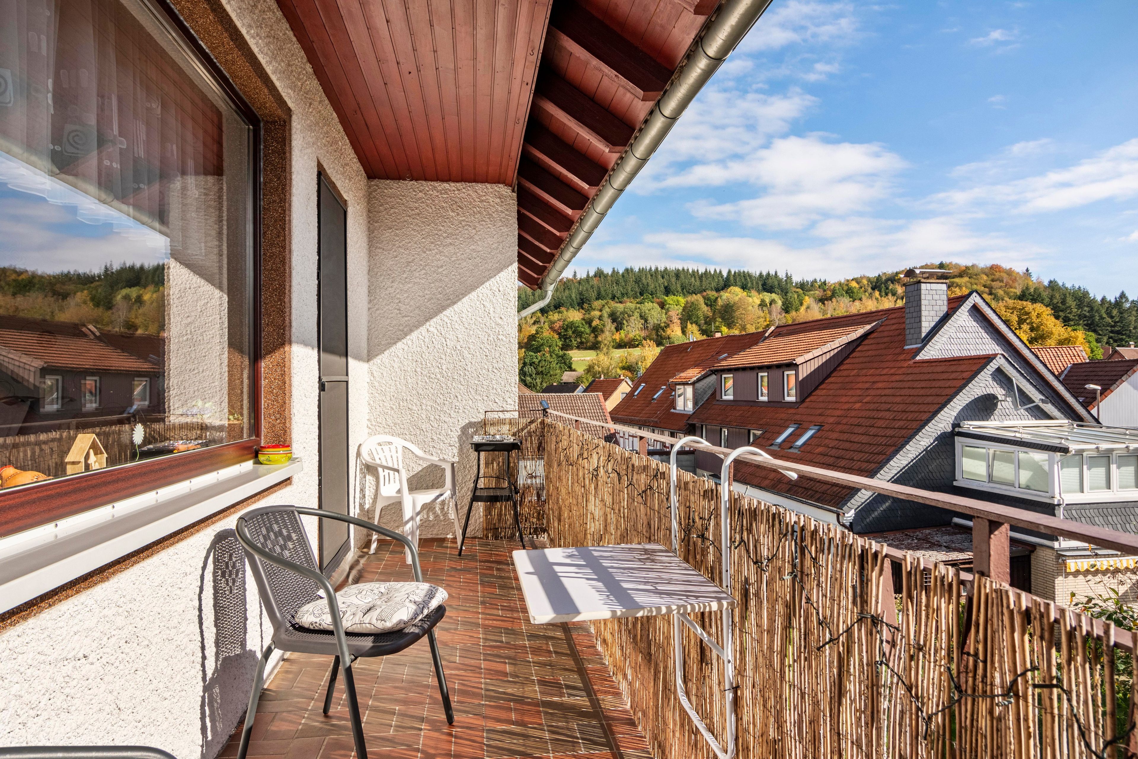 A balcony with a chair, a table, and a view of neighboring houses and a forested hill.