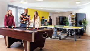 Een biljart met ballen, een tafeltennistafel en een automaat in een kamer met gele muren en een groot raam.