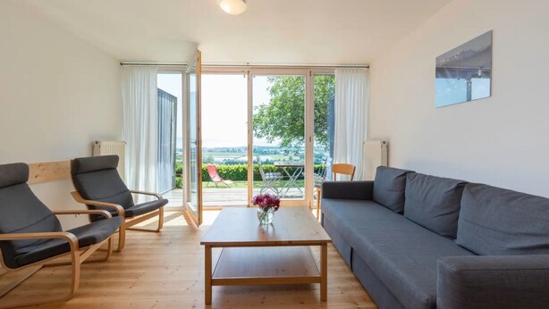 A modern living room with a sofa, two armchairs, a coffee table, and a view through large glass doors to an outdoor area with a table and chairs.