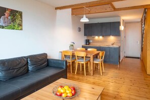 Un soggiorno moderno con un divano in pelle, un tavolo da pranzo con sedie, un angolo cottura con pensili scuri e un soffitto in legno, e una ciotola di frutta sul tavolino.