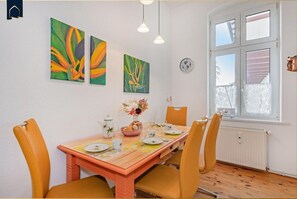 Un coin repas avec une table en bois, des chaises jaunes et une fenêtre avec des stores. Il y a trois tableaux au mur et une horloge.