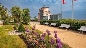 Un bâtiment doté d’une tour centrale, entouré d’un jardin aménagé avec des bancs et des fleurs violettes.