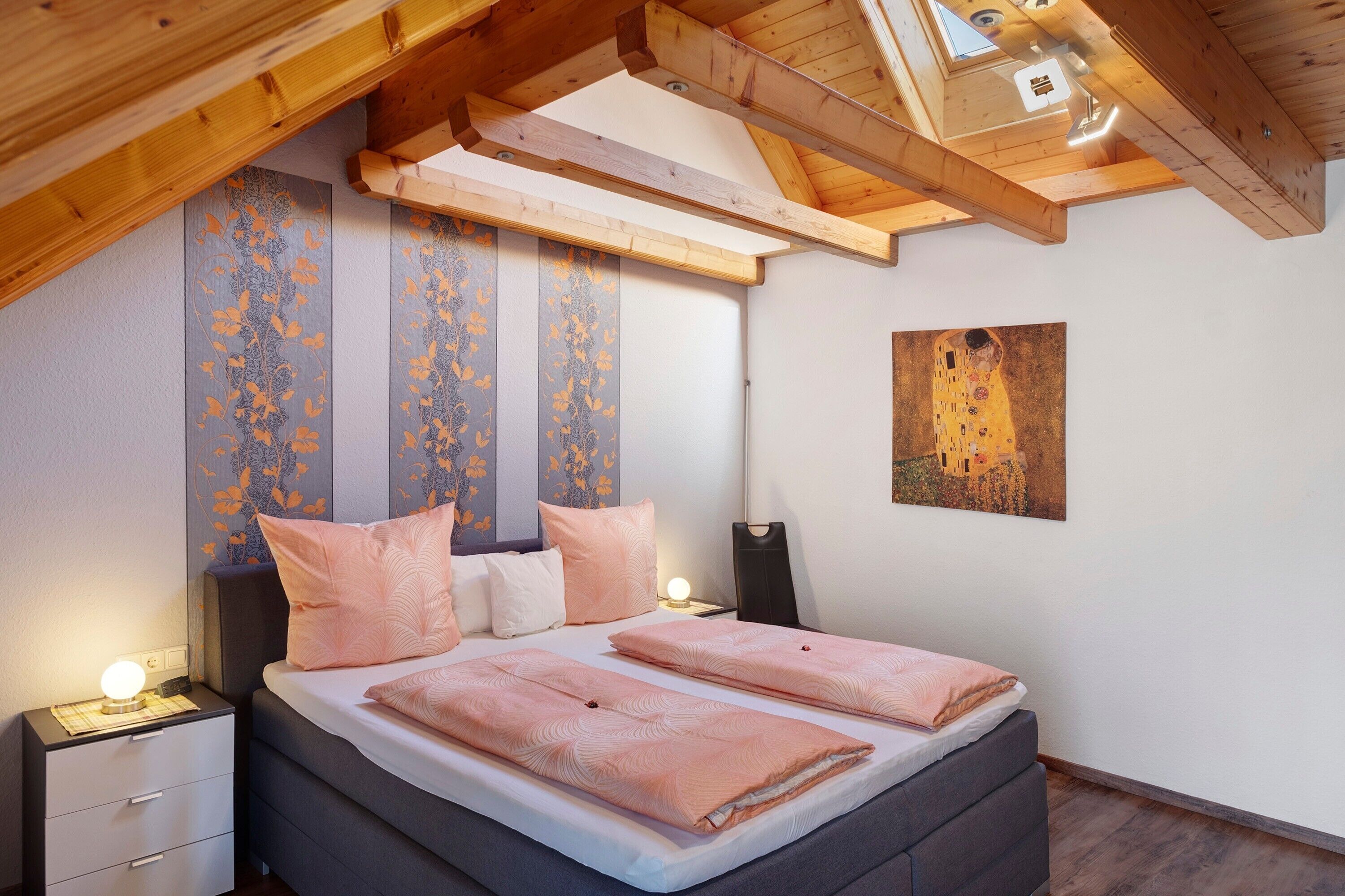 A bedroom with a bed, bedside tables, a nightstand, a ceiling with wooden beams, and a painting on the wall.