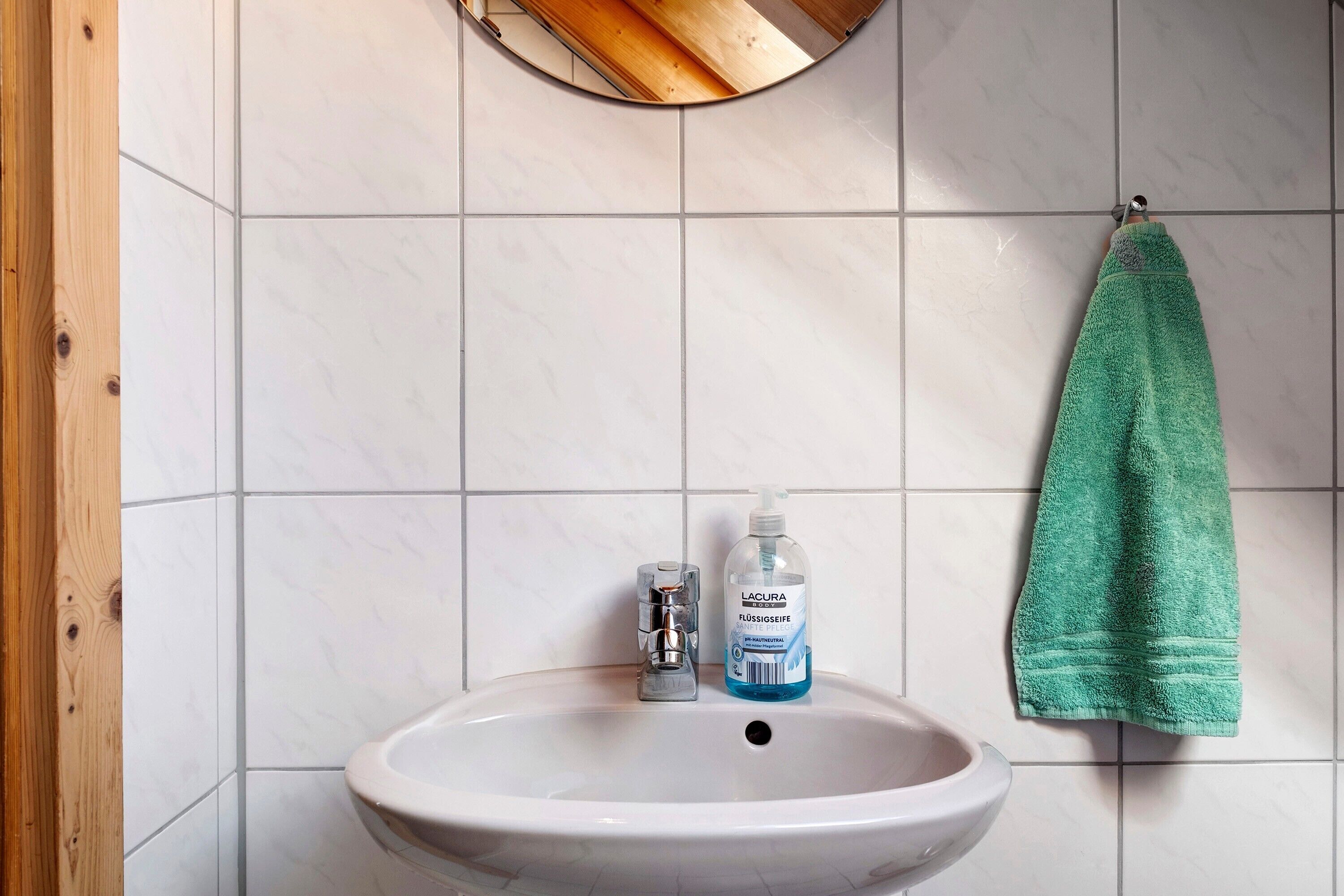 A bathroom with a white sink, a bottle of hand soap, and a green towel hanging on the wall.