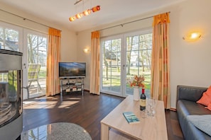 A modern living room with a flat-screen TV, a coffee table with a book and wine bottle, a sofa, and large windows with curtains. - "Ferien am Haff" Vacation Home with Sauna, Fireplace, and Terrace - Detached (Zirchow)
