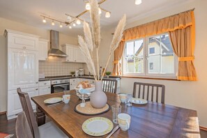 A dining area with a wooden table, chairs, a vase with pampas grass, and a window with curtains.