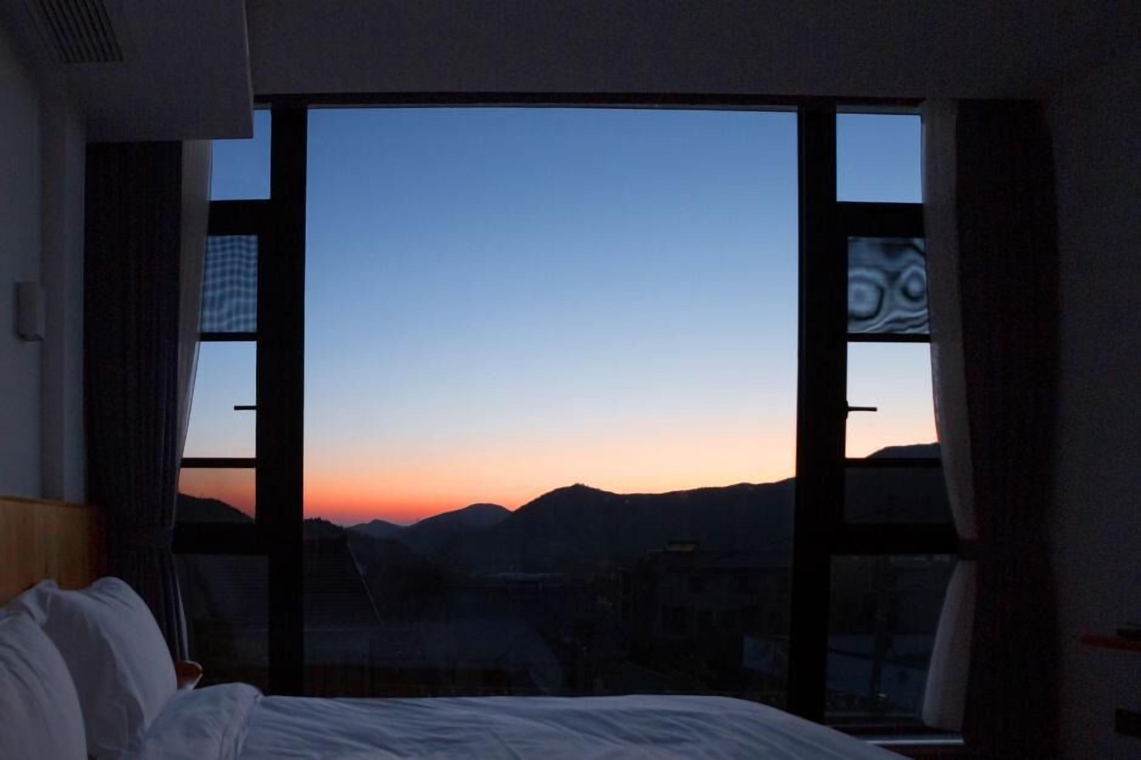 Habitación de hotel con un amplio ventanal que ofrece vistas de las montañas al atardecer.