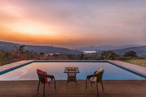 A pool with a view of mountains and a sunset. There are three chairs and a table with two candles.