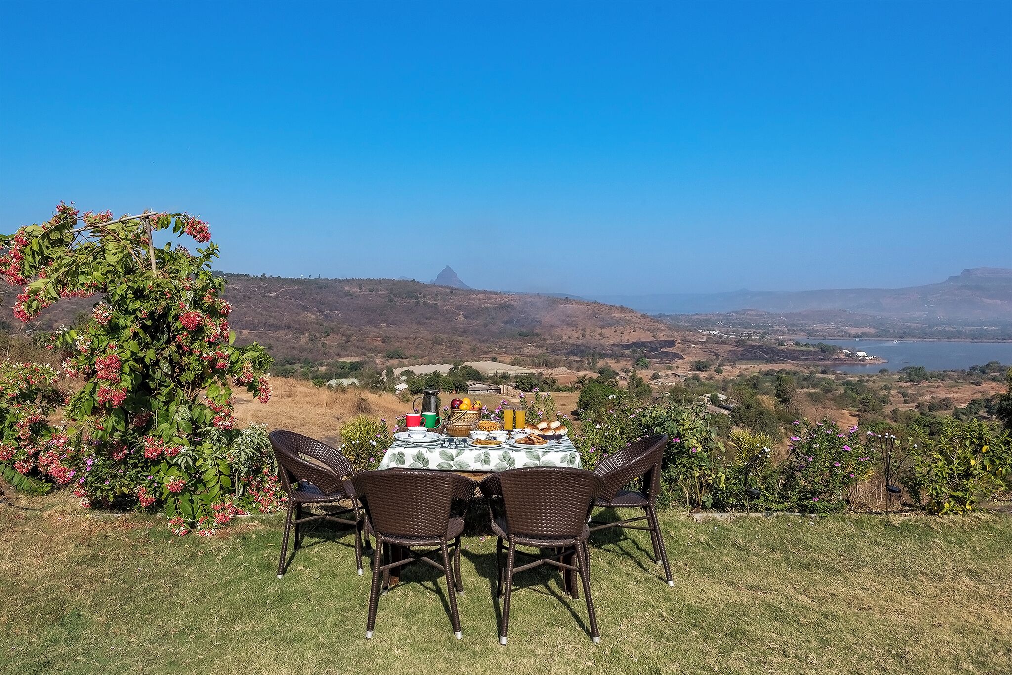 Un área de comedor al aire libre con sillas de mimbre y una mesa lista para una comida, con vistas a un paisaje pintoresco con colinas y una masa de agua.