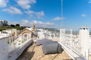 Una terraza en la azotea con baranda blanca, un edificio beige al fondo y un cielo azul despejado.