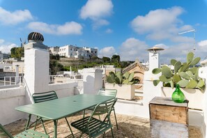 Una terraza en la azotea con una mesa y sillas de metal verde, una planta de cactus y vistas a edificios blancos al fondo.