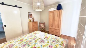 Quarto com cama, guarda-roupa de madeira, uma cadeira, luminária pendente e um quadro na parede.