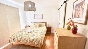 A bedroom with a wooden bed, a bedside table with a lamp, a hanging light fixture, a framed picture on the wall, and a small table with two vases.