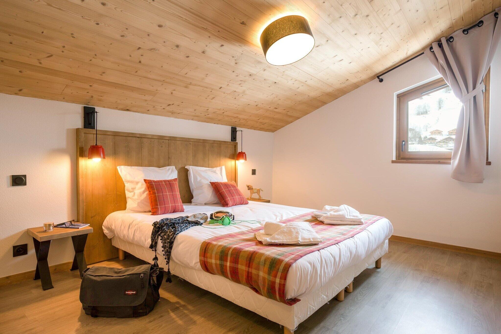 Un dormitorio con techo de madera, una cama con ropa blanca y una manta a cuadros, una mesita de noche con una lámpara y una ventana con cortinas.