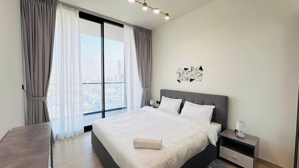 A modern bedroom with a large bed, a nightstand, a desk, and a view of the city through large windows. - Luxurious Belvilla 1BR JVC with Pool, Gym & BBQ Access (Dubai)