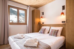 A double bed with two pillows, a wooden headboard, a window with curtains, and a wall-mounted lamp.