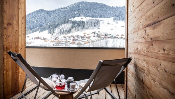 A wooden balcony with two chairs and a table, overlooking a snowy mountain village.