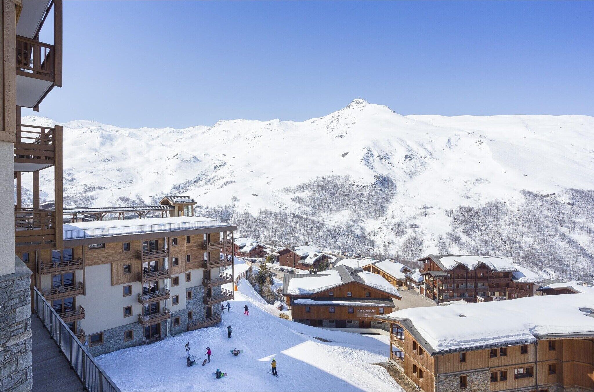 A snow-covered mountain resort with wooden buildings, ski slopes, and a clear blue sky.