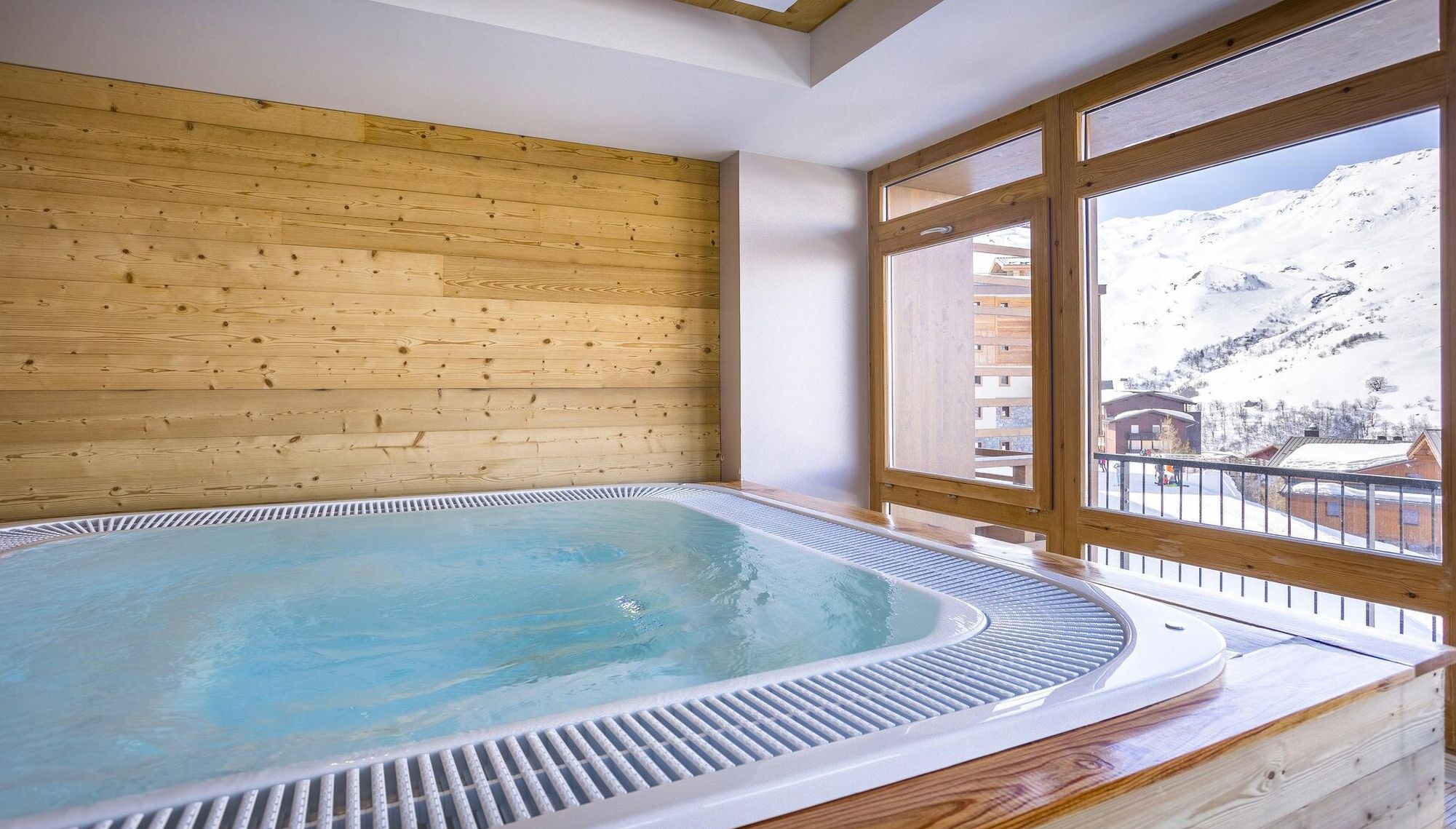 A hot tub in a room with wooden walls and large windows overlooking a snowy mountain.