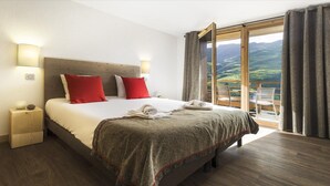 Ein Hotelzimmer mit einem großen Bett, zwei Nachttischlampen, einem Holzkopfteil und Blick auf eine gebirgige Landschaft durch eine Glastür.