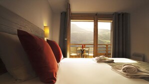 A hotel room with a bed, pillows, a table with chairs, a balcony with a table and chairs, and a view of the mountains.