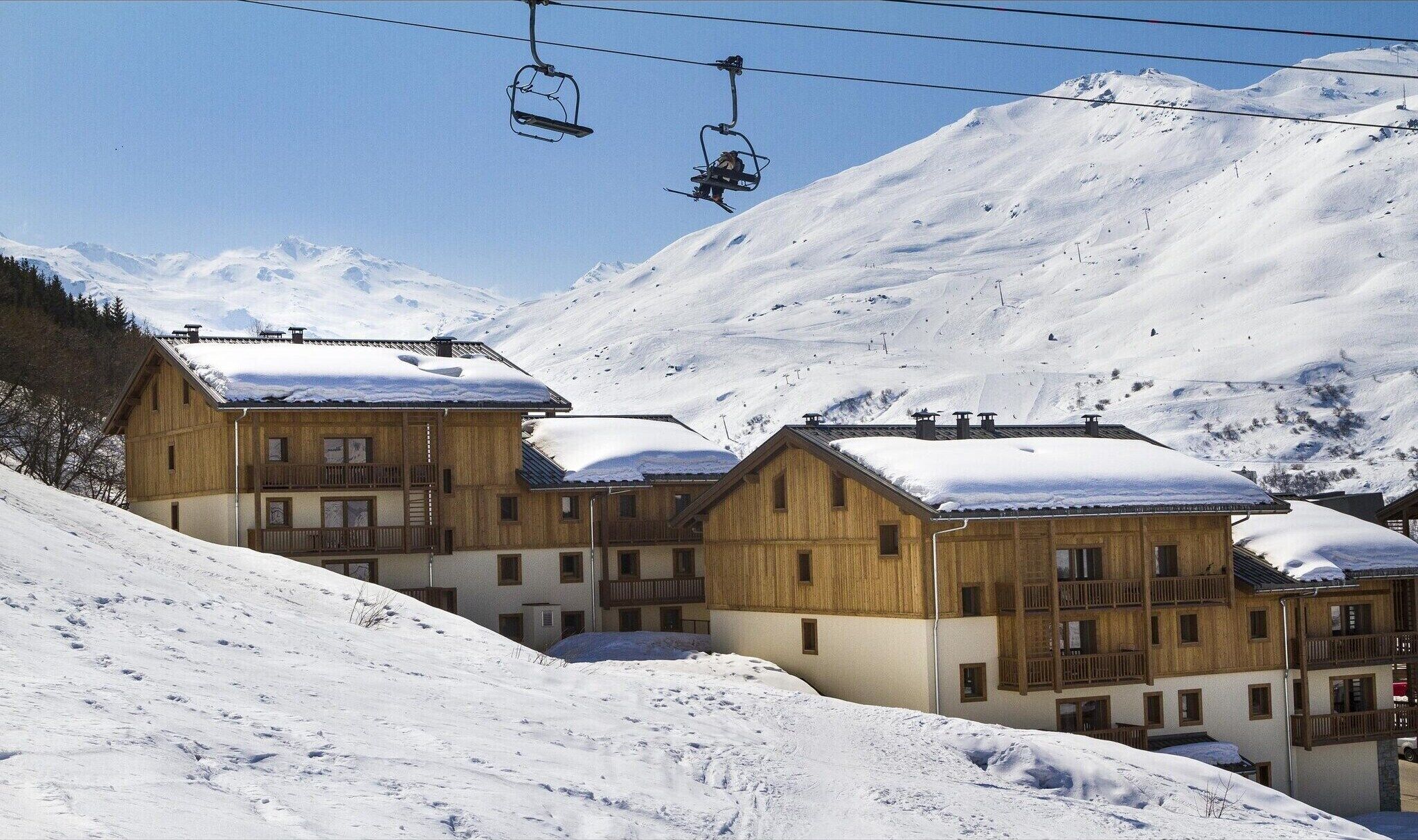 Een houten chalet met meerdere balkons, omringd door besneeuwde bergen en skiliften.